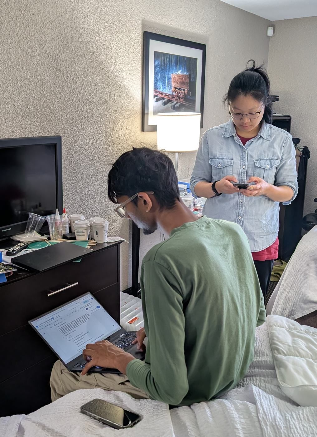 A member typing on a laptop in a hotel room while another checks a phone